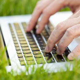 Close up image of a person typing on a laptop that is sitting in grass.